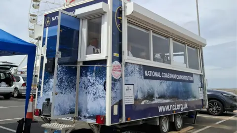 A mobile hut with three people inside. The exterior is blue and has a sign reading: NATIONAL COASTWATCH INSTITUTION