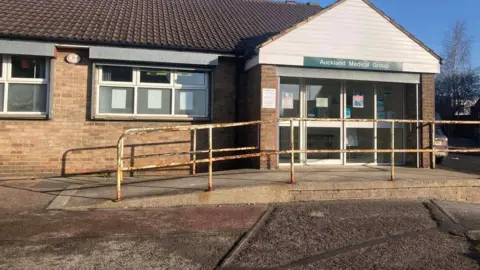 The outside of the surgery which is  one storey building with a rusty rail and large doors to the right of the picture. The building looks like it needs painting.