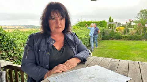 Juliet Jarvis with long dark hair wearing a blue jacket and sitting at a table on a lawn looking at a map. Two men are looking over a hedge behind her.