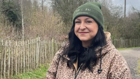 A woman wearing an animal print jacket with a park area behind her. She has long black hair and is wearing a dark green hat. 