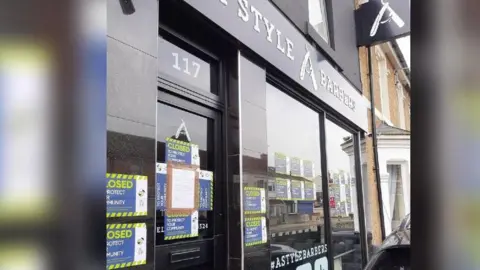 The exterior of the barbers in Swindon. It has around a dozen small 'closed' signs with police logos on the door and windows. The exterior is black and has large white lettering saying 'A Style Barbers'.