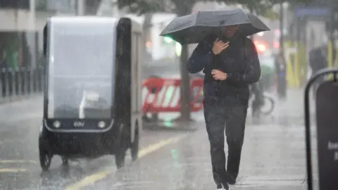 A man wearing a sodden jacket and trousers carries an umbrella down a street through a downpour 