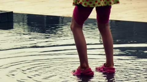 Getty Images A child wearing a green and pink polka dot dress, pink shorts and bright pink crocs, pictured splashing the fountain at Millennium Square. 