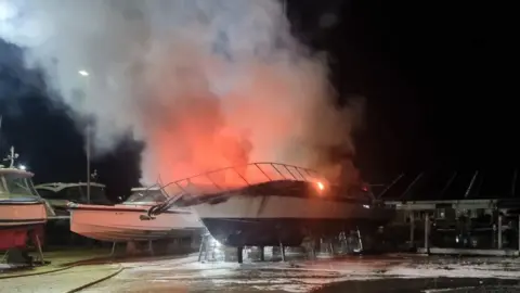 DWFRS Boat on a stand, appears to be a sailing boat, on fire in a boatyard.