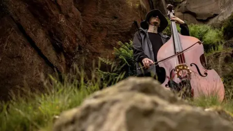 Bradford 2025 A double bass being played by a man in a raincoat and hat on Ilkley Moor