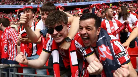 A stand of Sunderland fans - most wearing their team colours. Two youngs men in the foreground are raising their arms and shrieking with delight.