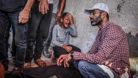 Anadolu caur Getty Images Bērns tup, tur galvu un raud blakus ķermenim zem melnas segas, kamēr vīrietis tup tuvumā, neizteiksmīgs, un blakus stāv citi vīrieši.