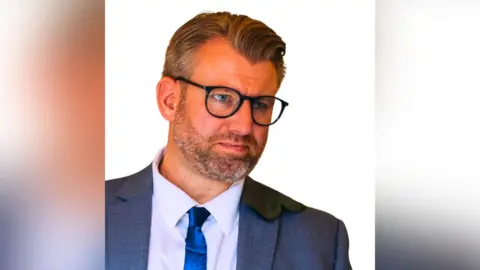 Security Industry Federation Daniel Garnham wears a grey suit, blue tie and white shirt. He has on black, rounded glasses and has a close cut beard. 