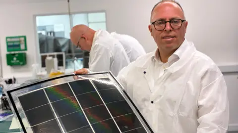 BBC/Richard Moss Neil Spann, who has short hair and is clean shaven with black framed glasses, hold a sheet of small black solar panels. He is weaeing a white shirt and lab coat. Behind him other people in white lab coats are bent over working on a table.