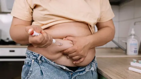An overweight woman lifting up a peach coloured t shirt above her blue jeans to inject a roll of fat on her stomach 