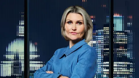 BBC/Freemantle A woman with shoulder length blonde hair and blue eyes is wearing a denim blue shirt and has her arms crossed and is looking at the camera. Behind her is a blurry scene of the London skyline.