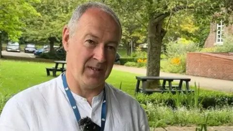 Nick Ireland is standing in a green space outside the Dorset Council offices. He has cropped, grey hair and is wearing a collarless shirt and a blue council lanyard.