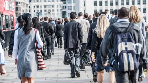 Lots of commuters walking along a pavement with their backs to the camera. Office buildings can be seen in the background