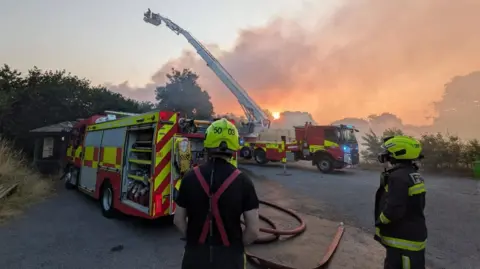 Two Devon & Somerset Fire & Rescue Service fire engines parked up outside the Occombe Farm site. There is a large amount of smoke in the sky. There are two fire fighters stood looking at the smoke. 