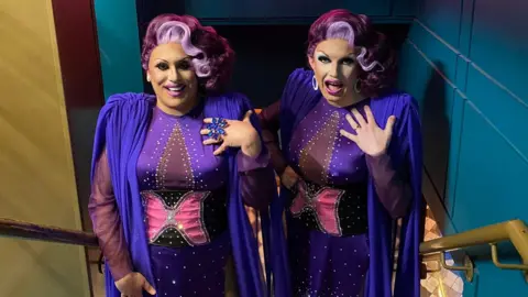 George and Jason Talbot-Sullovan standing on a staircase in matching purple and rhinestone drag costumes with pink and red wigs, full makeup and large jewelry. They are both wearing a blue/purple cape, style jacket with shoulder pads. Both are facing directly at the camera. 