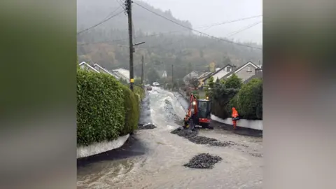 Samuel Guist A digger and a man in bright orange waterproof clothes are on a street with floodwater pouring down it. Trees on the side of mountains can be seen in the background.