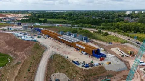 An aerial shot of a construction site next to a motorway, with green spaces and trees on the other side of the carriageway. A long brown structure is being built a few dozen feet from the motorway.