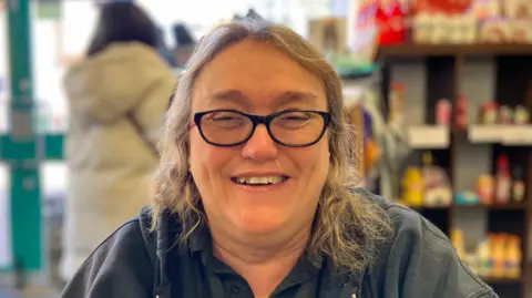 Lewis Adams/BBC A smiling Tina Townsend, who is wearing glasses with black rims and a navy polo top. She is sitting in the Witham Hub community centre, with a food shelf behind her.