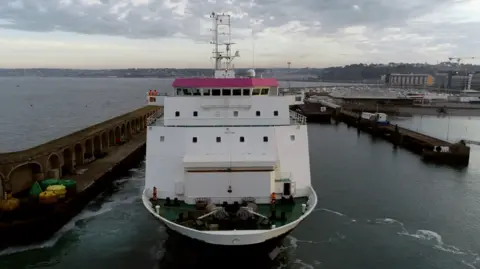 The front of the Commodore Goodwill with visual of the bridge and front deck. It is reversing into port at St Helier, with grey skies behind and the sea.