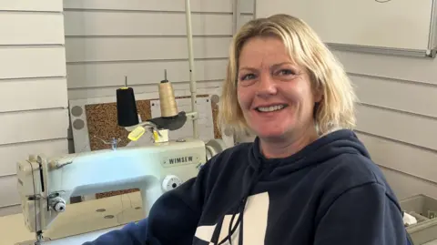 A blonde woman looks at the camera in a dark blue hooded top. A sewing machine can be seen in the background.