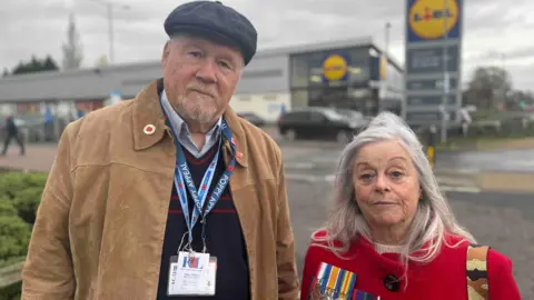 A man in a tan jacket and a black hat, wearing poppy badges stands next to a much shorter woman with mid length grey hair and a red coat. She's wearing a number of military medals as well as a red poppy. Both are in front of a blurred out Lidl supermarket. They're not smiling.