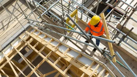 A bird's eye view of a builder wearing a hard hat and high visibility jacket works among trusses and scaffolding at a housing development.