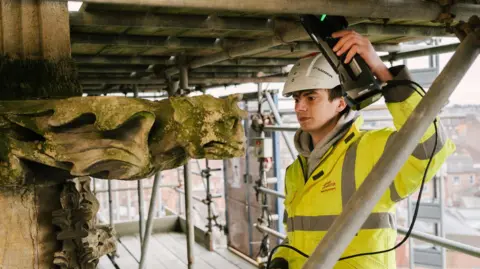 Luke wears a hi-vis jacket and hard hat, standing on scaffolding high up on the side of York Minster. He holds a 3D scanner in his left hand, pointing at a grotesque sculpture on the side of the wall.