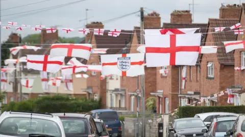 A photo of Torrington Avenue, a residential street in Knowle West, Bristol from 2016. There are houses all along the street and cars parked on both sides of the road. England flags have been put up all along the street.
