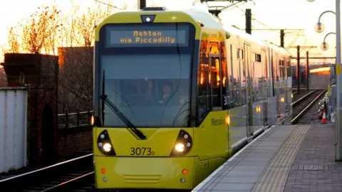 A yellow tram pulls into the station. It's taken on an early morning, with an orange sunrise seen in the distance.