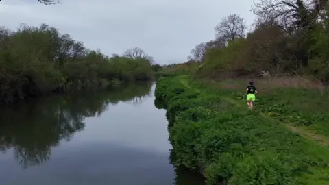 Heather Davey in the distance running along a riverbank, wearing green shorts and a black t-shirt.