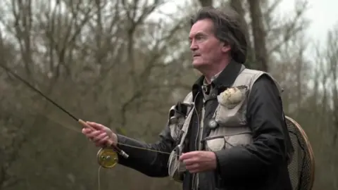 A man with dark brown hair who is fishing. He is holding the rod in his right hand and is holding the line in his left hand. He has a net on his back and wearing at sleeveless top which has lots of pockets. There are trees in the background.