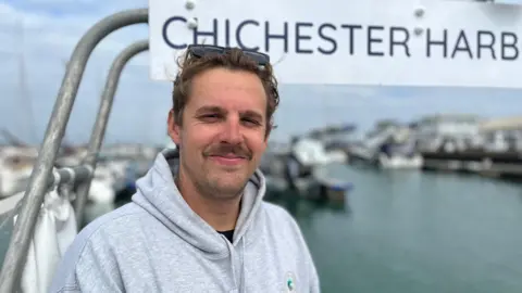 George Carden/BBC Patrick White standing on his boat which has the business logo on a metal frame above him. He has brown hair, a grey jumper and sunglasses.