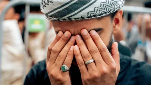 PA Media A man wearing a black and white headdress and holding his hands against his eyes with two rings on his fingers.
