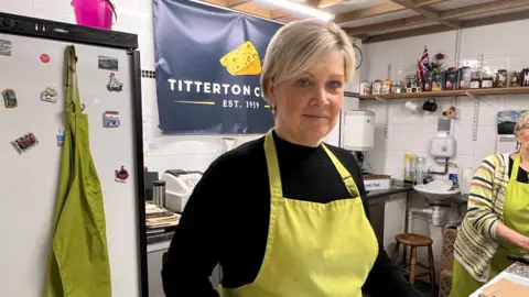 BBC Sharon wears a black tp and yellow apron, has short blonde hair and is standing behing a cheese counter. 