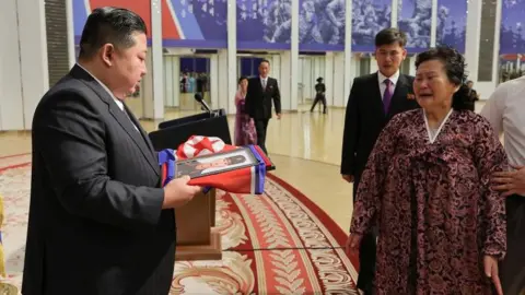 Kim Jong Un, wearing a dark suit, presents a crying woman with a photo draped in a blue and red flag. The woman is wearing a dark red dress and they are in a grand hall. 