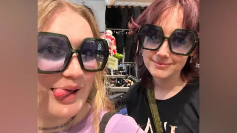 Phoebe Ashley Two university students - one with long blonde hair and another with shoulder-length purple hair - pose in a shop as they try on sunglasses.