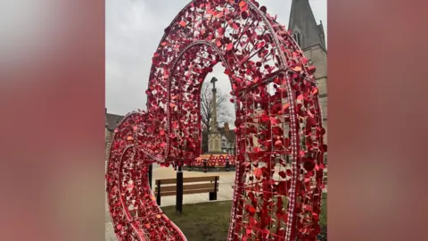 The heart-shaped seat. It is red. A church can be seen in the background where there are also wreaths around a memorial. 