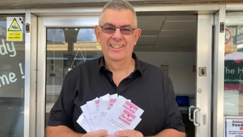 A man wearing a black shirt is standing in front of an office building, holding up several sheets of paper which have ideas written on them. 