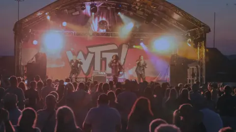 The festival in Melksham in 2025. A three-piece musical group, two in sparkly black clothes and one in a sparkly pink outfit, performing on a stage at night time. Behind them is a large sign reading "WTF" – referring to Wiltshire Throwback Festival. Outlines of audience members are in the foreground.