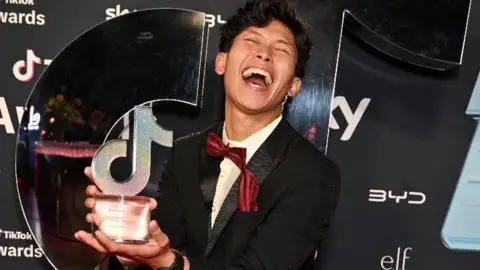 Gab Astorga, who has black wavy hair, closes his eyes, throws his head back and laughs , while holding a glass and mirror TikTok award. He is dressed in a black dinner suit with red bow tie and is standing in front of a black branded screen.
