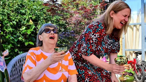 Handout A woman with short grey hair, an orange and purple print top and sunglasses sits in a garden chair and grins while holding a glass of champagne. Perched on the arm of the chair is another woman, with longer light brown hair and wearing a red, black and blue print dress, she is laughing and has her eyes closed.