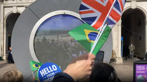 Andrew Woodger/BBC A circular "portal" showing an image of a crowd of people on a beach. People in front of the portal hold Brazilian and Union Jack flags