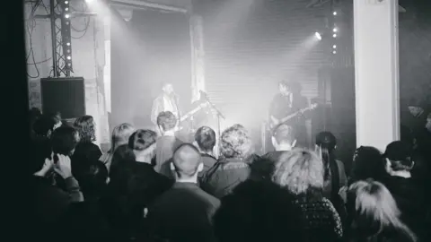 A live show inside The White Hotel venue. Two men are playing guitars on stage and a crowd watch.