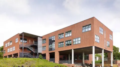 Modern buildings at Moseley School with children in uniform outside in the playground. The building is on columns with an open area underneath and grassy areas around it.