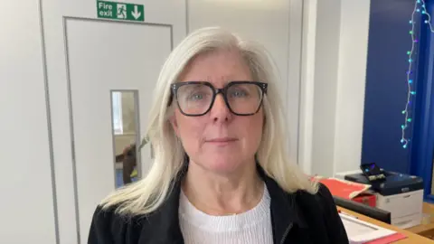 A close-up of a woman with shoulder-length silver hair, wearing a black jacket, white shirt and dark-rimmed glasses.