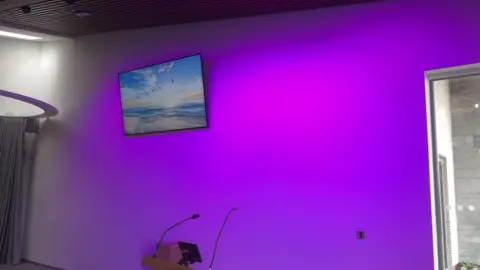 A small TV screen is hung on a wall to the right of the photo which is displaying a beach scene with sea and a sunset above it with birds flying across. To the left of the photo a bright pink and purple light shines in a white wall. There is also a lectern with a microphone in the middle of the photo