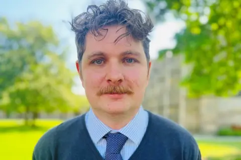 Dan Cutts, Aberdeen co-chair of the UCU, a man with wavy dark brown hair, a moustache, and a blue shirt, tie, and jumper.