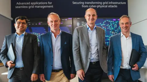 Four representatives of Siemens and Camlin Group walking towards the camera alongside one another. They are smiling and looking at the camera. All four are dressed in jackets and shirts of different colours - mostly greys and blues.