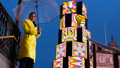 Art of London Lakwena Maciver is seen in a bright yellow coat holds a clear umbrella beside a tall, illuminated, multi-coloured cube sculpture at dusk, with historic buildings in the background.