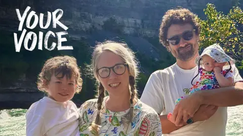 A smiling woman with two long blonde plaits in her hair and wearing glasses and a long sleeved knee length summer dress with a tropical print, stands on some fenced decking over a green rushing river. She is holding a smiling toddler, a boy with brown wavy hair and wearing a white t-shirt, colourful shorts and blue sandals. Next to her is a taller man with curly brown hair wearing sunglasses, a white t-shirt and red shorts and black trainers, he is smiling and holding a small baby in a flowery summer romper suit and a white sunhat. 